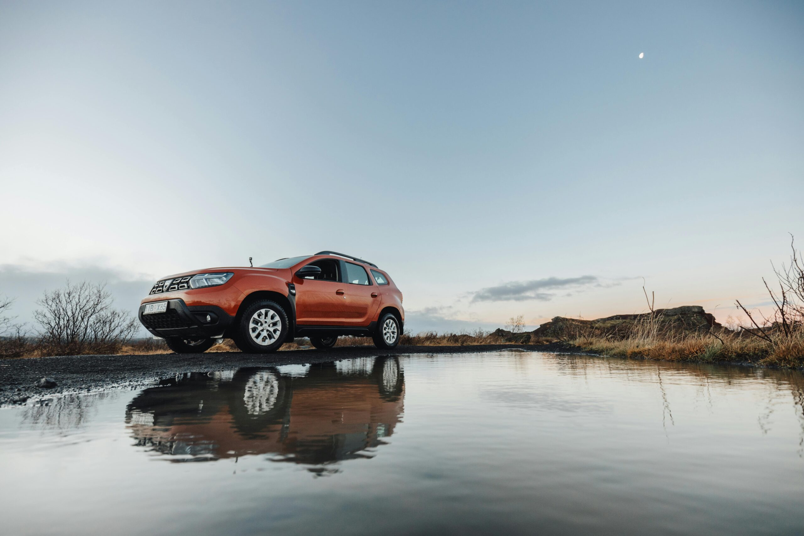 Home A vibrant orange SUV parked on a road after rain, with a puddle reflecting the vehicle against a serene dusk sky.
