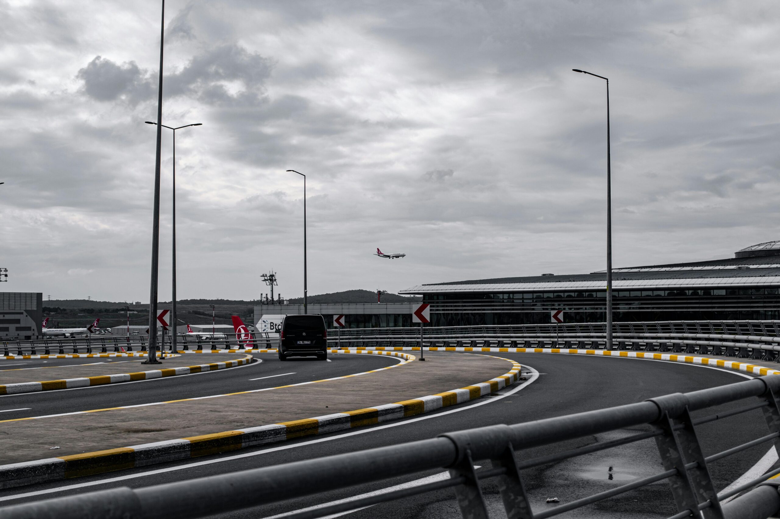 Home An airplane approaches for landing near a modern airport terminal with a curving road in the foreground.
